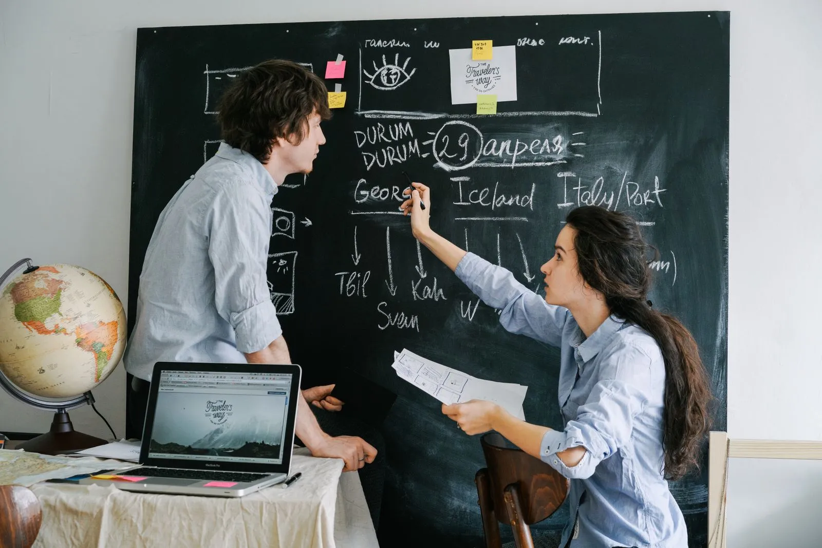 Two people planning on a chalkboard next to a laptop and a globe, like a host mapping out a direct booking plan