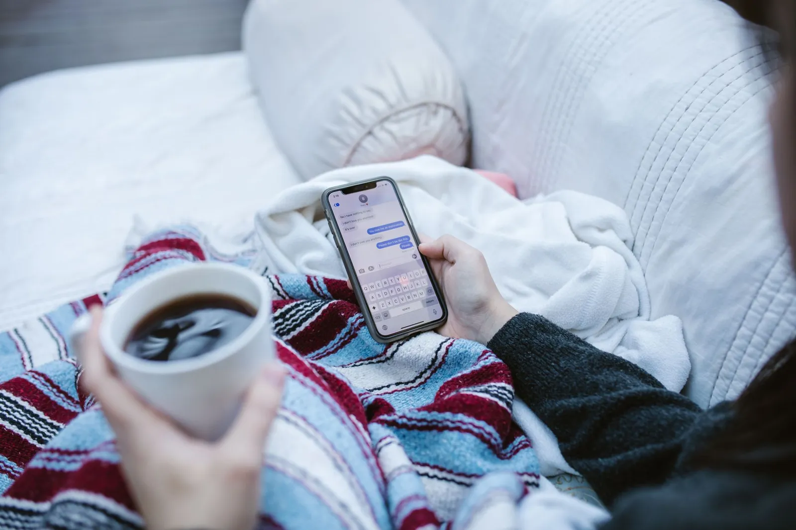 Persona escribiendo en un smartphone con un café, envuelta en una manta
